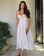 Woman in a white floral dress standing in a sunlit room with a decorative gate and plant.