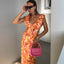 Woman in an orange and red patterned dress holding a pink handbag indoors.