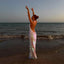 Woman in a sparkly dress standing on a beach with arms raised at sunset.