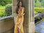 Woman in a yellow dress standing in a garden with stone arches and greenery.